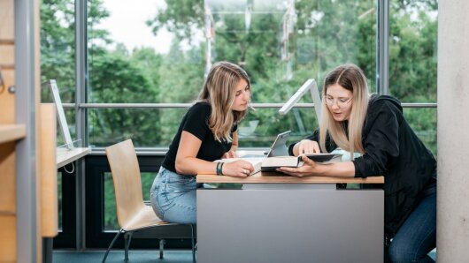 Zwei Studentinnen sitzen in der Bibliothek und schauen gemeinsam in ein Buch.